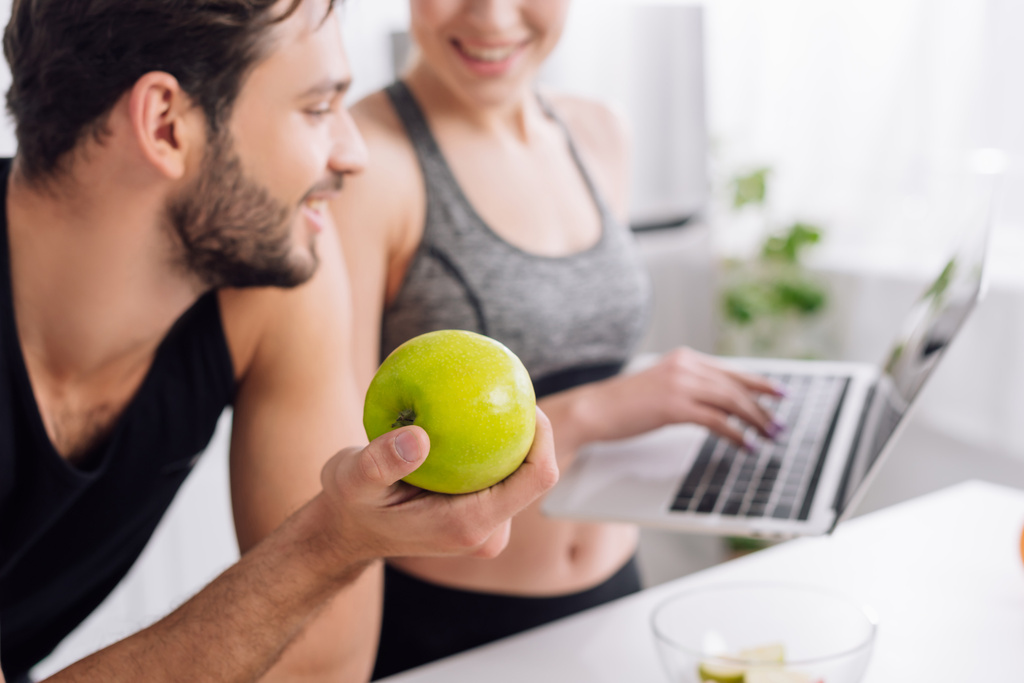 selectieve focus van gelukkig man houden appel en kijken naar laptop in de buurt van vrouw  - Foto, afbeelding