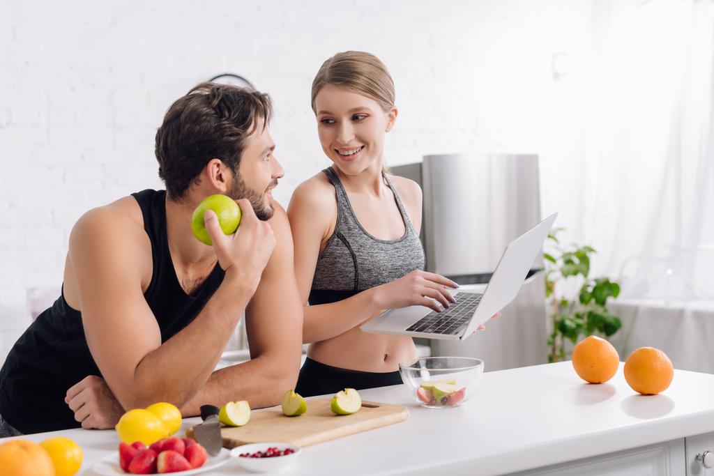 ευτυχισμένος άνθρωπος με μήλο κοιτάζοντας γυναίκα με φορητό υπολογιστή κοντά σε φρούτα  - Φωτογραφία, εικόνα