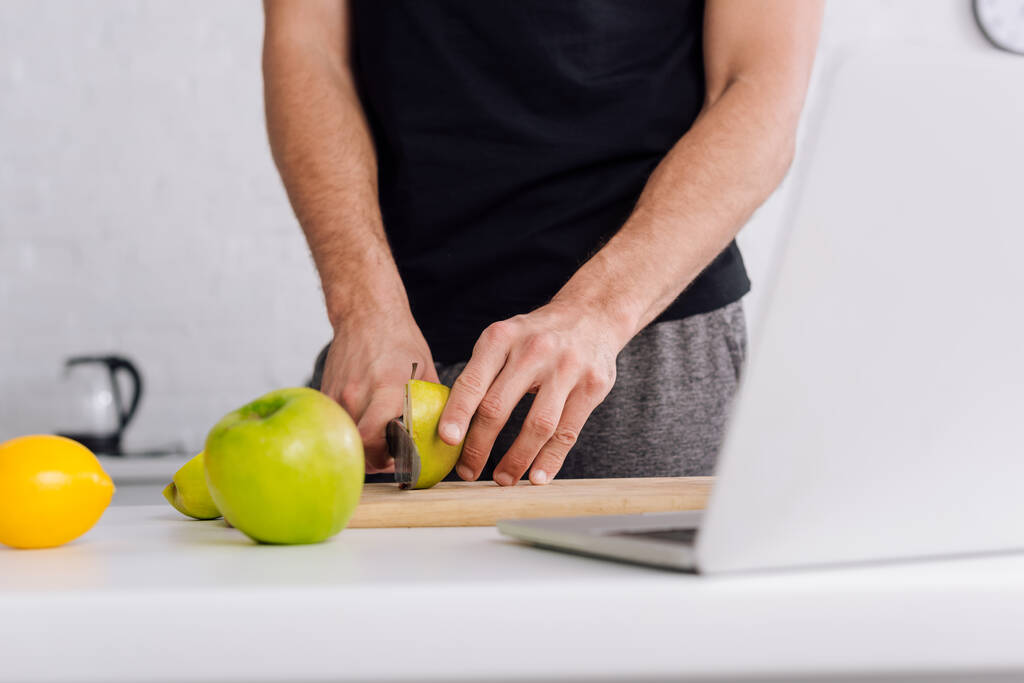 bijgesneden beeld van de mens snijden appel in de buurt laptop thuis - Foto, afbeelding
