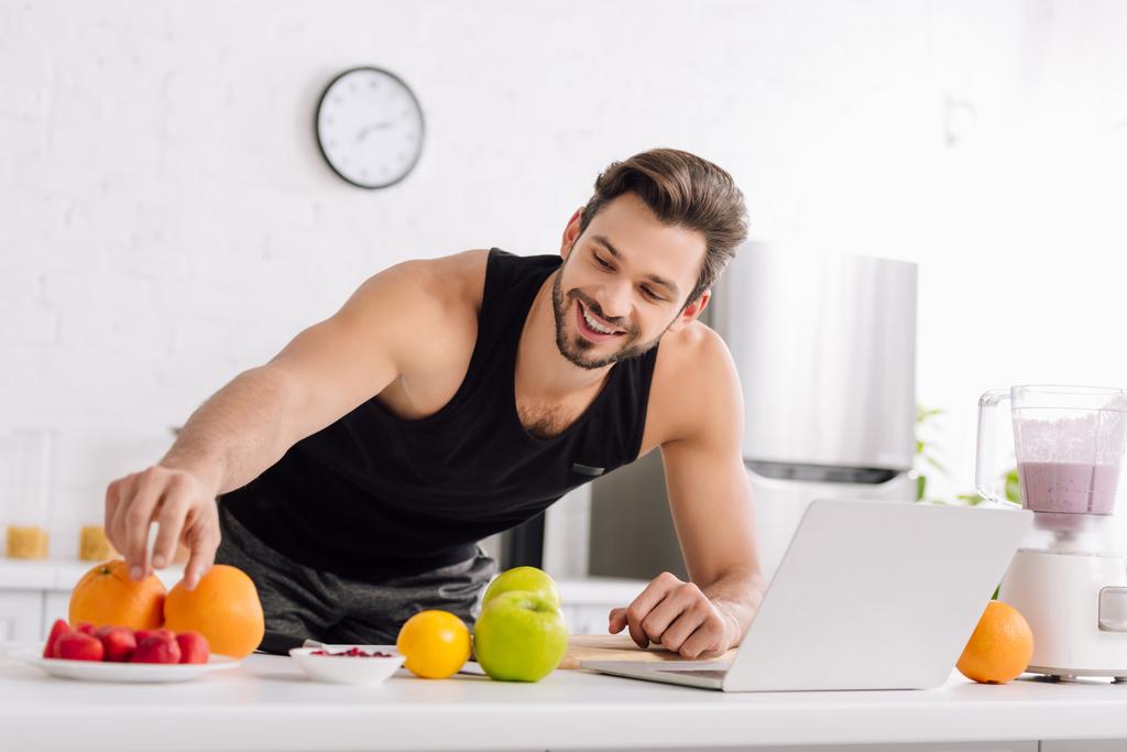 gelukkig man in de buurt van laptop, blender met smoothie en lekker fruit  - Foto, afbeelding