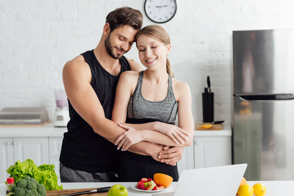 felice uomo abbracciando ragazza in abbigliamento sportivo vicino laptop e frutta fresca
  - Foto, immagini