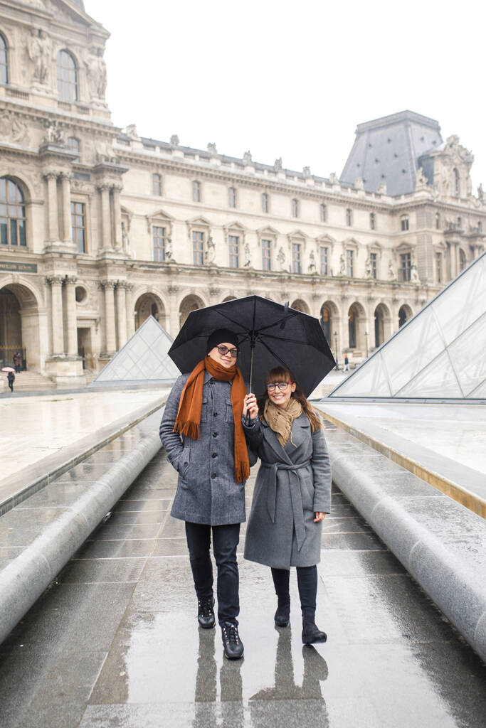 パリのルーブル美術館の近くには、若い美しいカップルがスタイリッシュに身を包み、灰色のコートの女の子、灰色のコートの男が歩いています。 - 写真・画像
