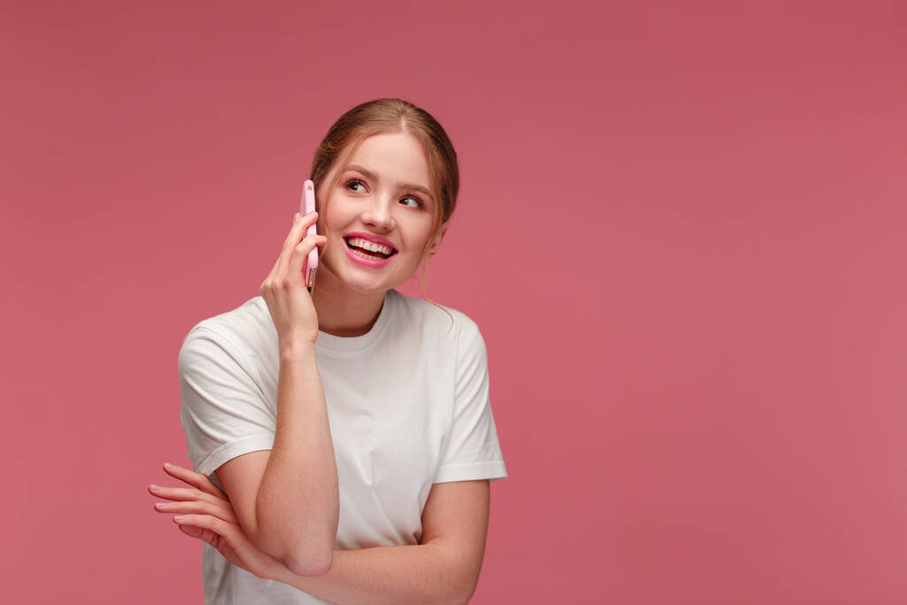 携帯電話で話している白いTシャツを着たかわいい幸せな赤毛の女の子の肖像画とピンクの背景に隔離された笑顔。コピースペース。モバイル機器を扱う若者は - 写真・画像