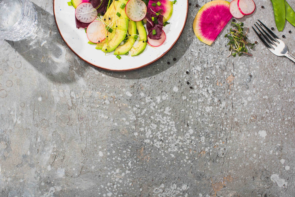 Draufsicht auf frischen Rettichsalat mit Gemüse und Avocado auf grauer Betonoberfläche mit Glas Wasser und Gabel - Foto, Bild