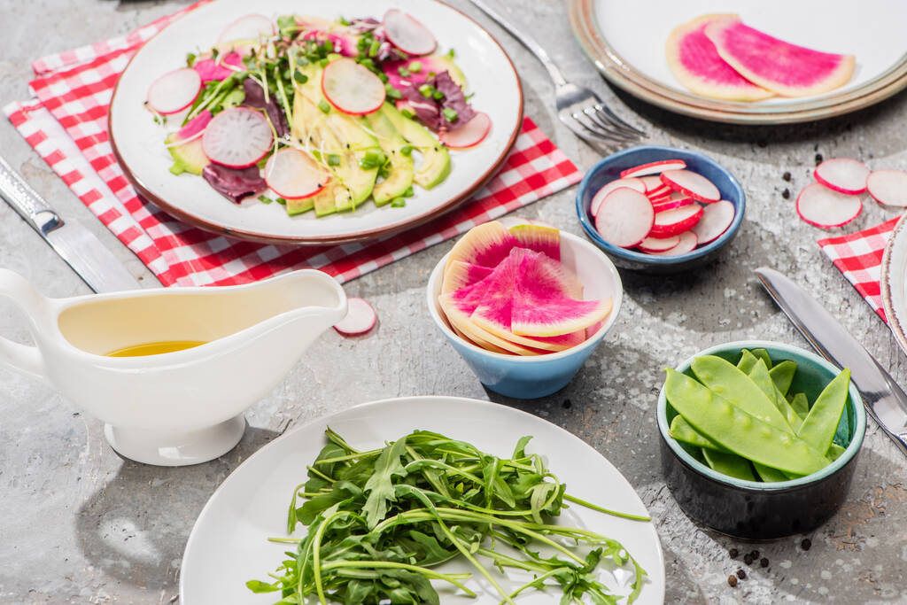 verse radijssalade met groenten en avocado in bord geserveerd op servet met bestek in de buurt van ingrediënten in kommen op grijs betonnen oppervlak - Foto, afbeelding