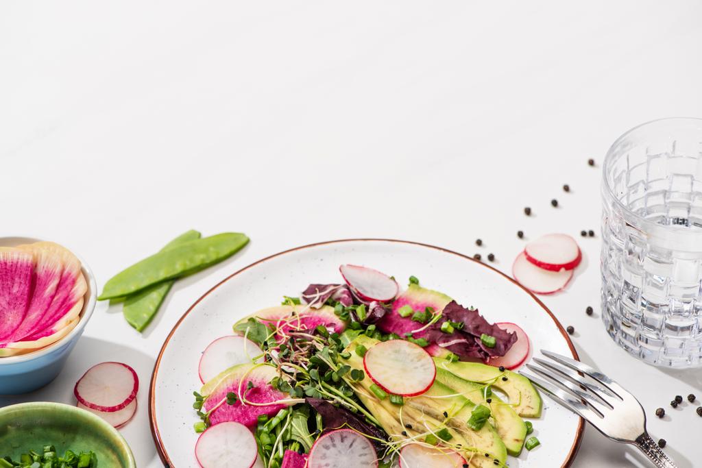 čerstvý ředkvičkový salát se zelení a avokádem na bílém povrchu se složkami v miskách, vodě a vidličce - Fotografie, Obrázek