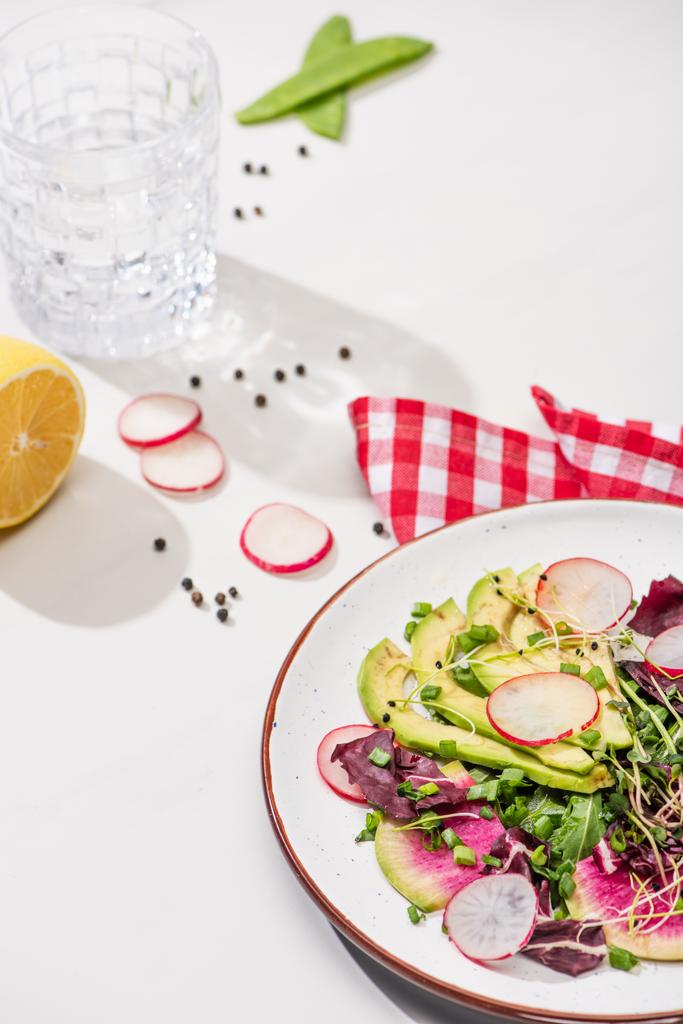 selektiver Schwerpunkt von frischem Rettichsalat mit Gemüse und Avocado auf Teller auf weißer Oberfläche mit Wasser, Zitrone und Serviette - Foto, Bild