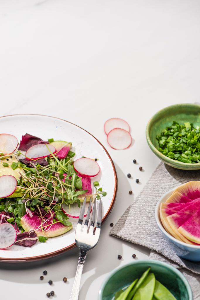 свіжий салат з редькою з зеленню та авокадо на тарілці на білій поверхні з інгредієнтами у мисці та виделці
 - Фото, зображення