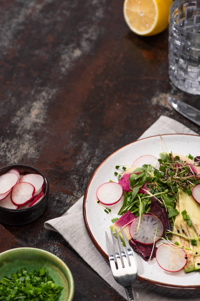 verse radijssalade met groenten en avocado op bord geserveerd op verweerd oppervlak met bestek, citroen en water - Foto, afbeelding