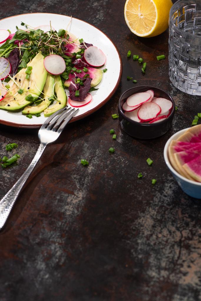 verse radijssalade met groenten en avocado op plaat op verweerd oppervlak met vork, citroen en water - Foto, afbeelding