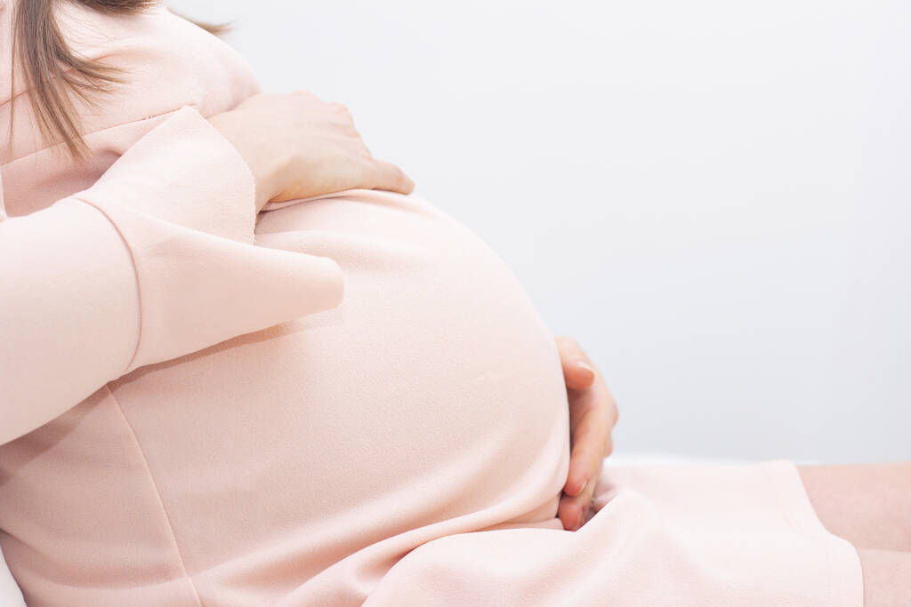 terhes nő ruhában fogja a kezét a hasán, könnyű háttérrel. Terhesség, anyaság, felkészülés és elvárás koncepció. Közelkép bent. Gyönyörű gyengéd hangulatú fotó terhesség. - Fotó, kép