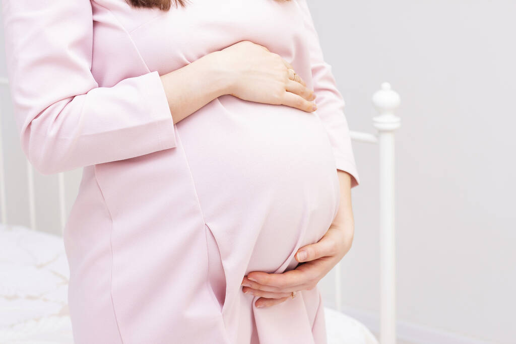 terhes nő ruhában fogja a kezét a hasán, könnyű háttérrel. Terhesség, anyaság, felkészülés és elvárás koncepció. Közelkép bent. Gyönyörű gyengéd hangulatú fotó terhesség. - Fotó, kép
