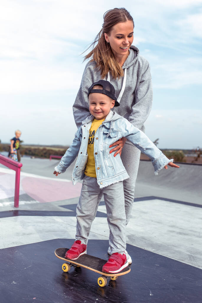 Woman mom, holds little boy son 3-5 years old, learning to ride a skateboard, in the summer on the sports ground, emotions of happiness, fun, relaxation and pleasure. Casual wear. - Fotoğraf, Görsel