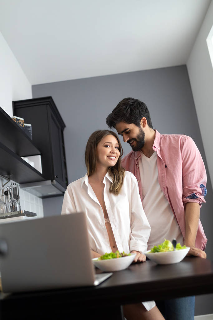 Молодая пара разговаривает и улыбается, готовя здоровую еду на кухне дома - Фото, изображение