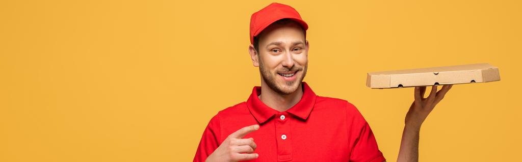 šťastný doručovatel v červené uniformě ukazuje prstem na krabici od pizzy izolované na žluté, panoramatický výstřel - Fotografie, Obrázek