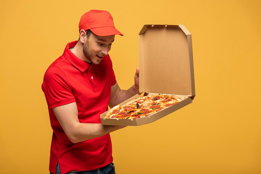 boldog futár piros egyenruhában nézi doboz finom pizza elszigetelt sárga - Fotó, kép