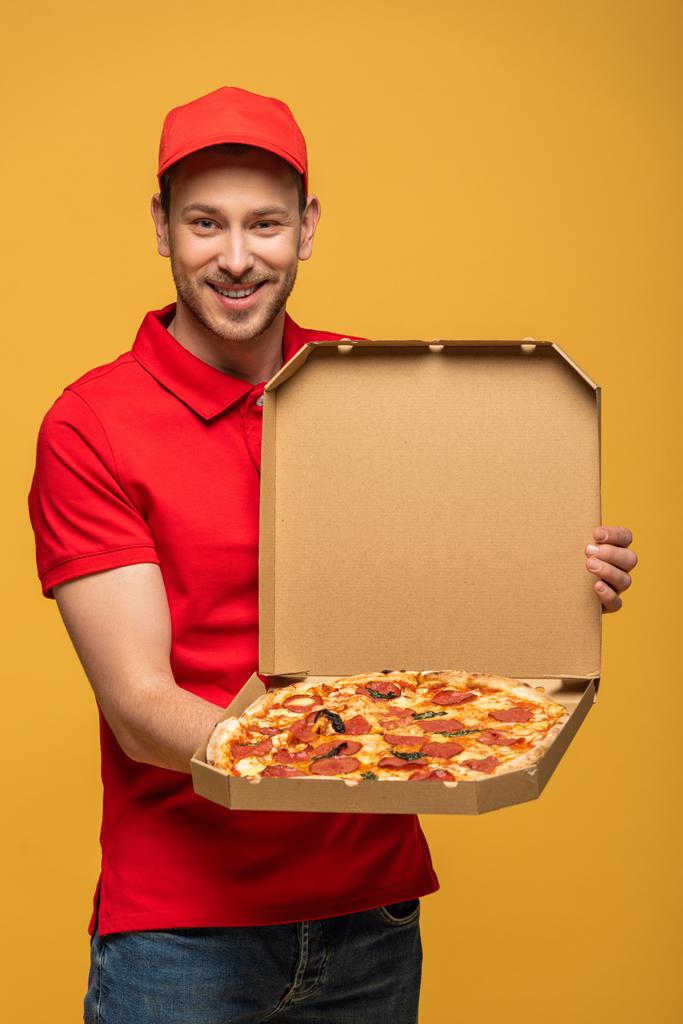 ευτυχισμένος άνθρωπος παράδοση σε κόκκινο στολή δείχνει κουτί με νόστιμα πίτσα απομονώνονται σε κίτρινο - Φωτογραφία, εικόνα