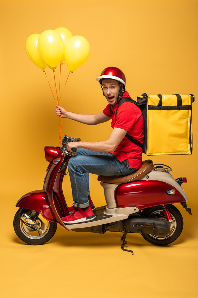вид сбоку возбужденного доставщика с воздушными шарами и рюкзаком на скутере на желтом фоне
 - Фото, изображение