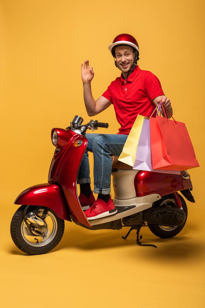 uśmiechnięty dostawca w czerwonym mundurze z torbami na zakupy machający ręką na skuterze na żółtym tle - Zdjęcie, obraz