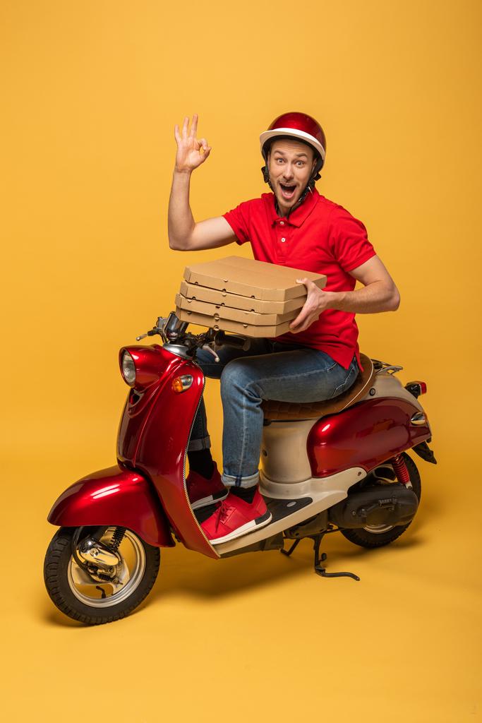 взволнованный курьер в красной форме держит коробки пиццы и показывает ОК знак на скутере на желтом фоне
 - Фото, изображение