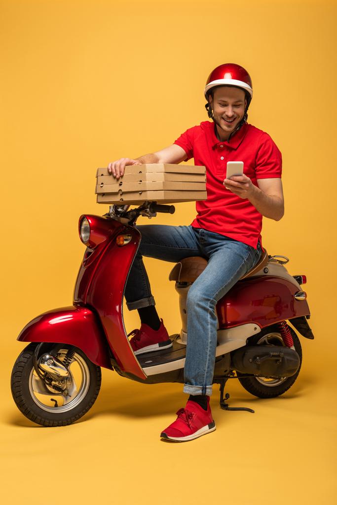 Kırmızı üniformalı mutlu teslimatçı elinde pizza kutuları ve sarı arka planda scooter 'da akıllı telefon kullanıyor. - Fotoğraf, Görsel