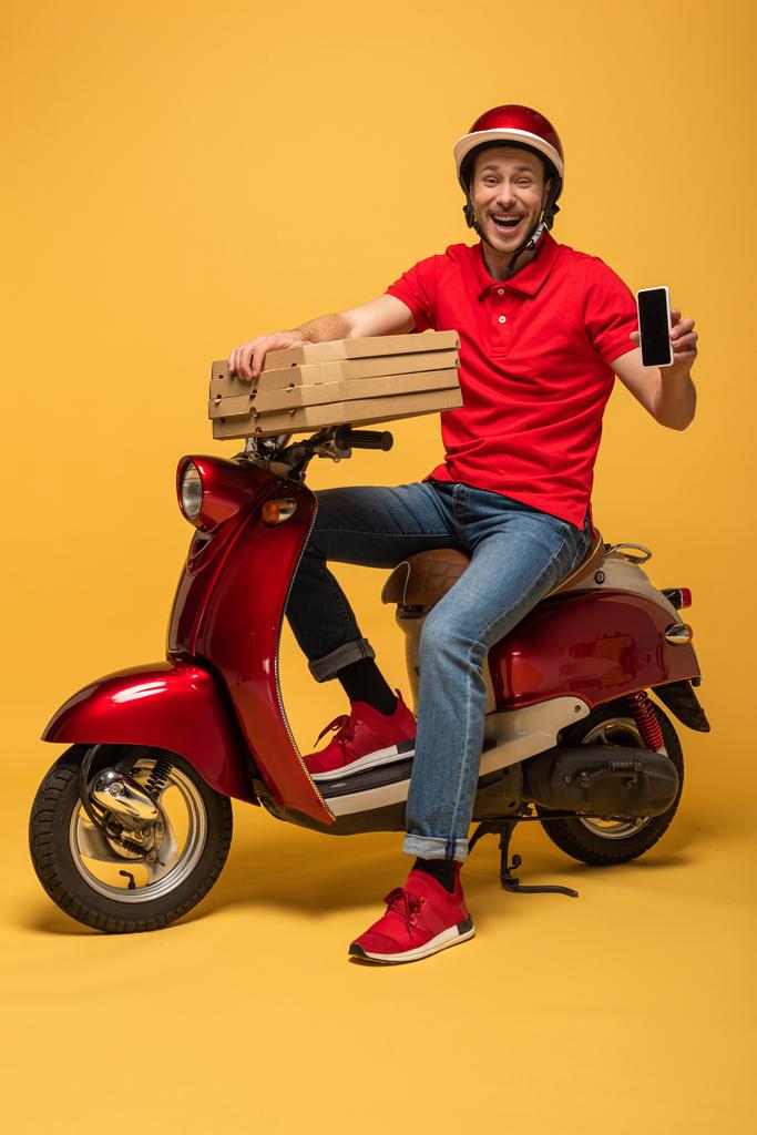счастливый курьер в красной форме держит коробки пиццы и смартфон с пустым экраном на скутере на желтом фоне
 - Фото, изображение