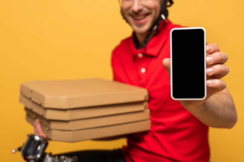 赤い制服を着た幸せな配達人のクロップドビュー黄色に隔離された空白の画面を持つピザボックスとスマートフォンを保持 - 写真・画像