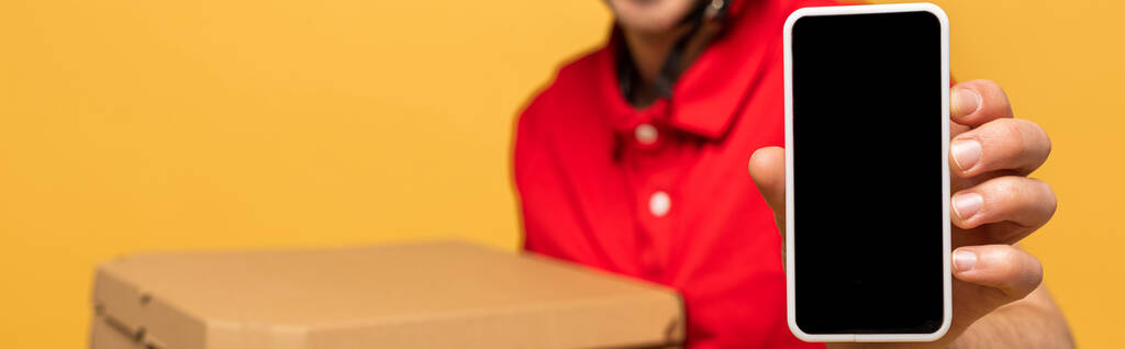 Kırmızı üniformalı mutlu teslimatçının elinde pizza kutuları ve sarı panoramik çekimde izole edilmiş boş ekranlı akıllı telefon vardı. - Fotoğraf, Görsel