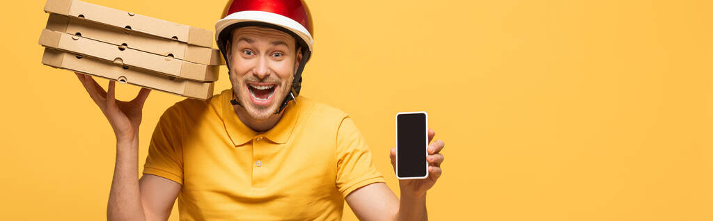 黄色の制服を着た興奮した配達人黄色の画面で隔離されたスマートフォンを示すピザボックスパノラマショット - 写真・画像