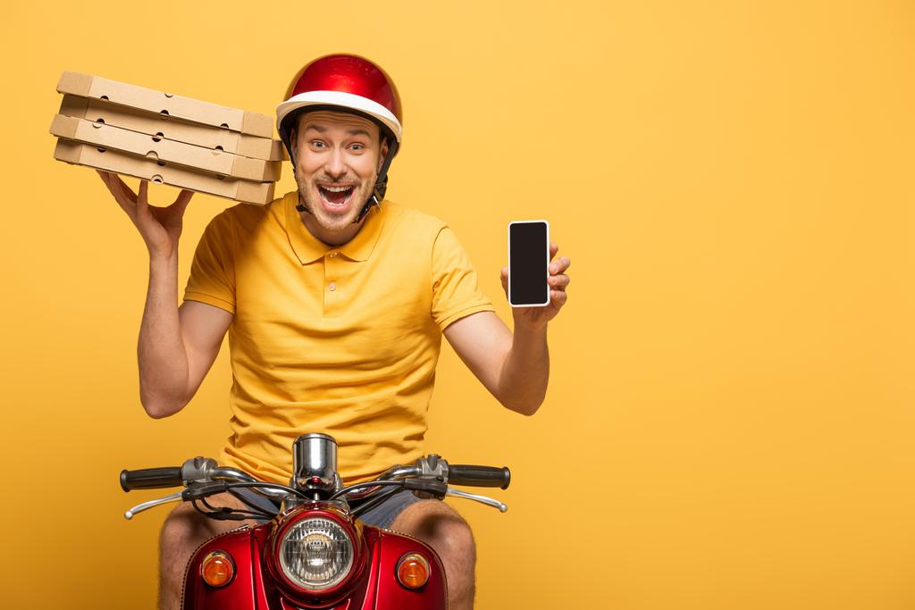 ενθουσιασμένοι delivery man σε κίτρινη στολή ιππασίας σκούτερ με κουτιά πίτσα και δείχνει smartphone με λευκή οθόνη απομονώνονται σε κίτρινο - Φωτογραφία, εικόνα
