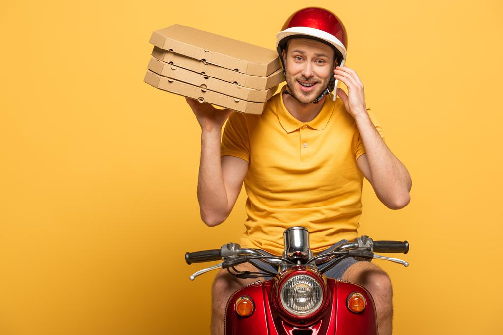 repartidor sonriente en scooter uniforme amarillo con cajas de pizza y hablando en smartphone aislado en amarillo
 - Foto, Imagen