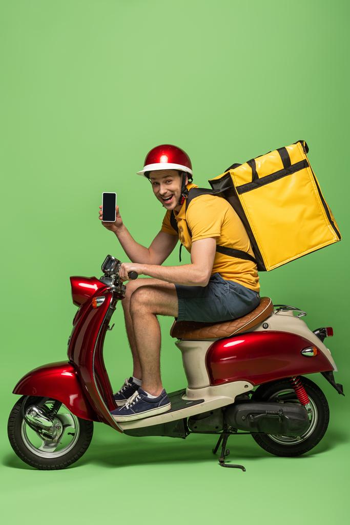 Вид сбоку счастливого доставщика в желтой форме с рюкзаком, показывающим смартфон с блатным экраном на скутере на зеленом
 - Фото, изображение