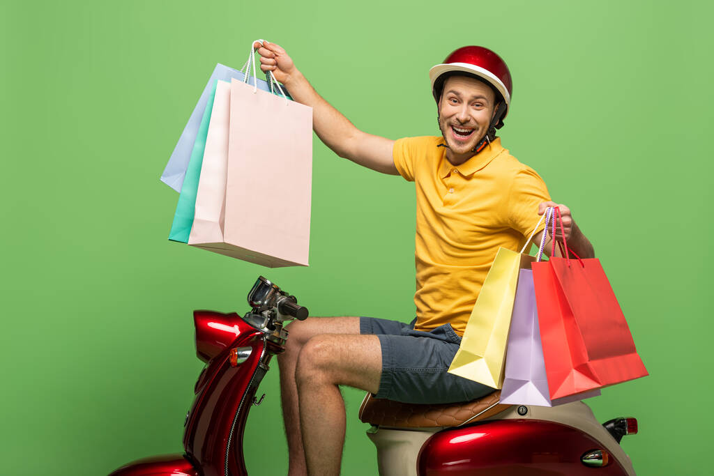 vista lateral do homem de entrega sorridente em uniforme amarelo e capacete segurando sacos de compras em scooter no verde
 - Foto, Imagem