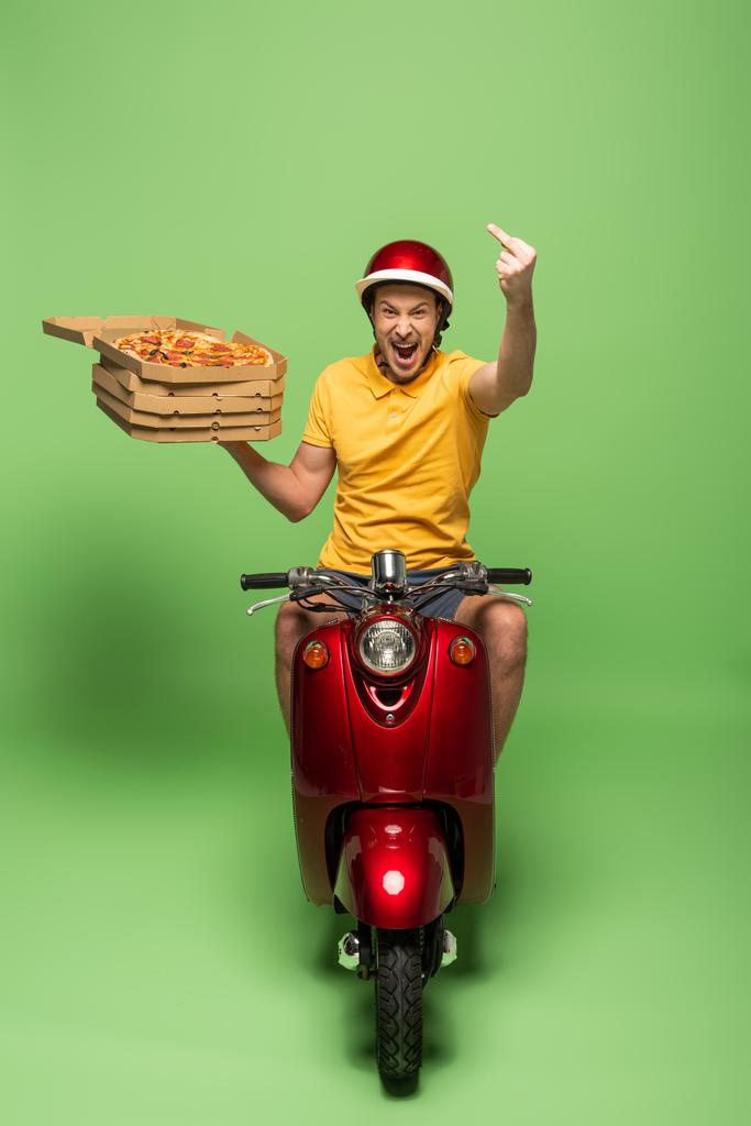 opgewonden levering man in geel uniform met open mond op scooter leveren pizza en tonen middelvinger op groen - Foto, afbeelding