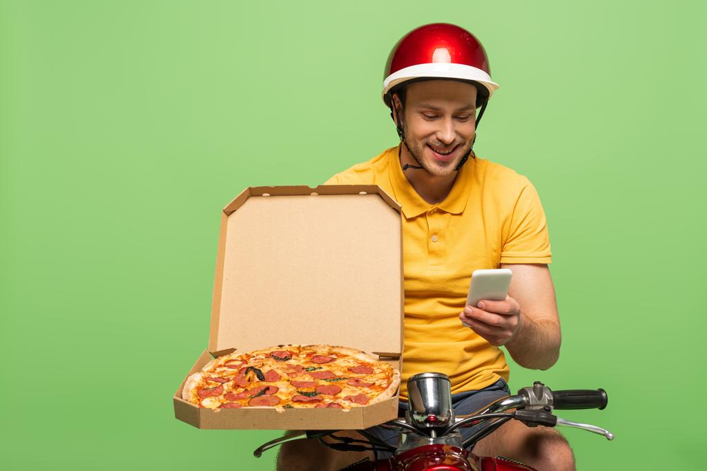 улыбающийся курьер в желтой форме на скутере с пиццей, используя смартфон, изолированный на зеленом
 - Фото, изображение