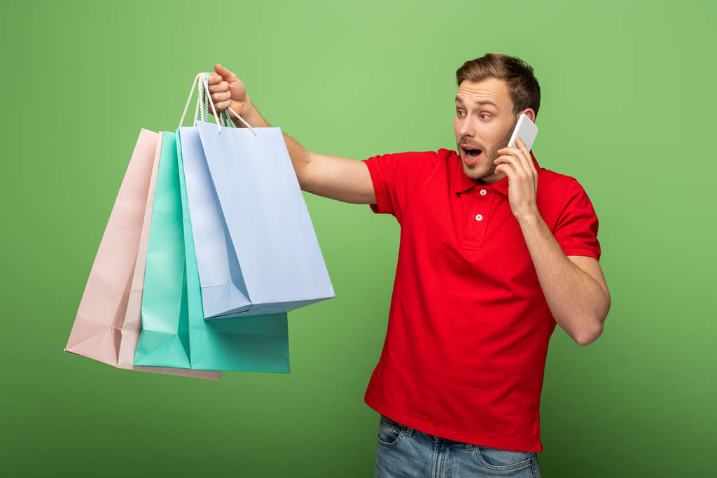 σοκαρισμένος άνθρωπος κρατώντας τσάντες ψώνια, ενώ μιλάμε για smartphone στο πράσινο - Φωτογραφία, εικόνα