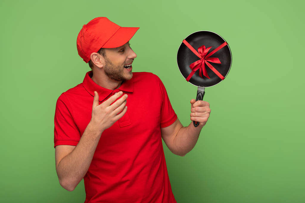 happy delivery man in red uniform holding frying pan with bow on green - Photo, Image