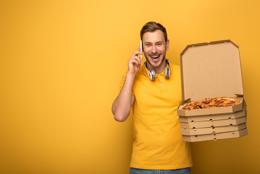 ευτυχισμένος άνθρωπος με ακουστικά σε κίτρινο στολή κρατώντας πίτσα και μιλώντας στο smartphone σε κίτρινο φόντο - Φωτογραφία, εικόνα