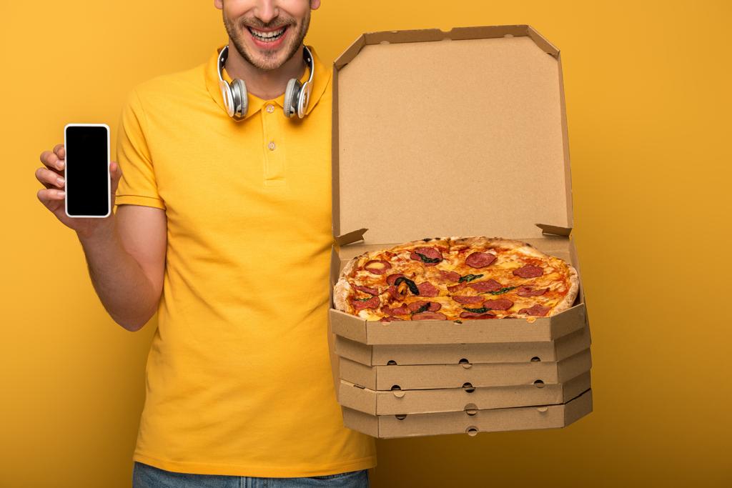Ausgeschnittene Ansicht eines glücklichen Mannes mit Kopfhörern in gelbem Outfit, der Pizza und Smartphone auf gelbem Hintergrund hält - Foto, Bild