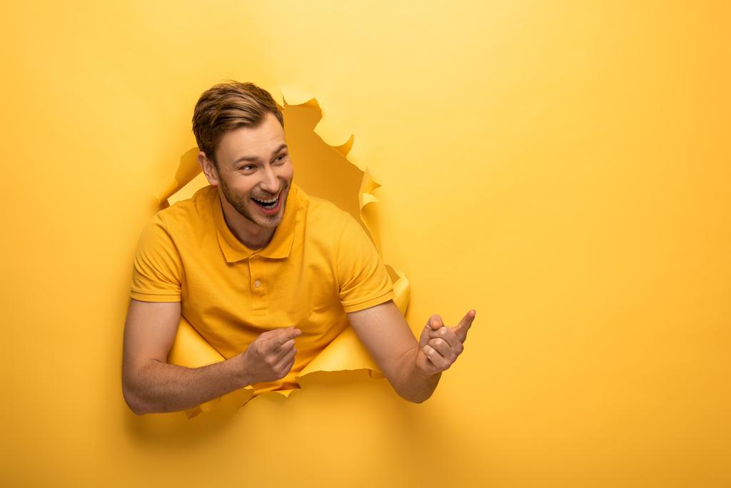 happy handsome man in yellow outfit in yellow paper hole pointing with fingers aside - Photo, Image