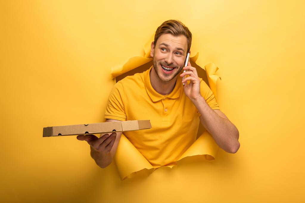 glücklicher gutaussehender Mann in gelbem Outfit, der mit dem Smartphone spricht und eine Pizzakiste im gelben Papierloch hält  - Foto, Bild