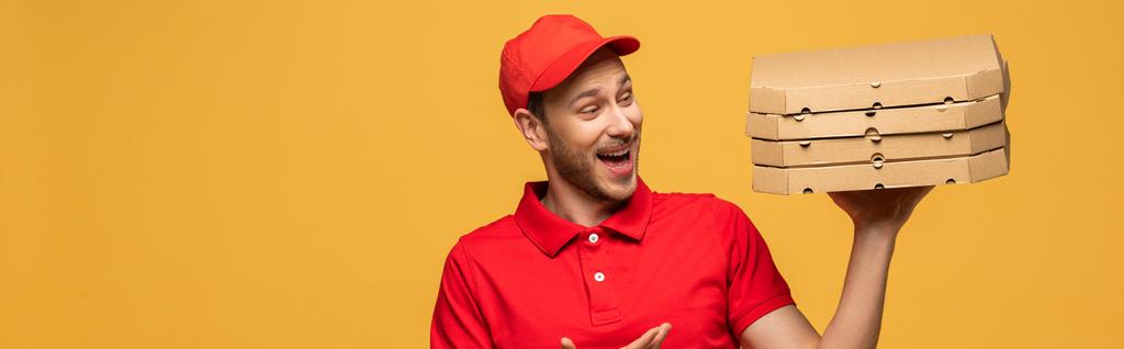 glücklicher Zusteller in roter Uniform, der mit der Hand auf Pizzakartons zeigt, isoliert auf gelbem Grund, Panoramaaufnahme - Foto, Bild