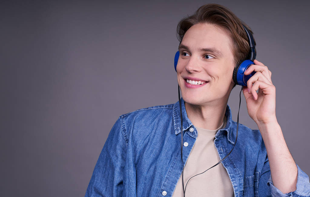 Leuke knappe jongeman, heel gelukkig, luisterend naar de soundtrack in blauwe koptelefoon, met een denim shirt, naar links kijkend, geïsoleerd op een grijze achtergrond. Kopieerruimte. - Foto, afbeelding