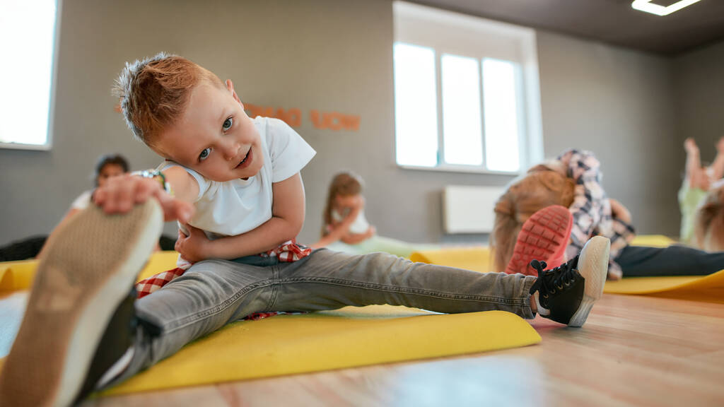 Flexibility. Close up portrait of a little boy sitting on yoga mat and stretching his legs. Group of children doing gymnastic exercises in the dance studio - Fotografie, Obrázek