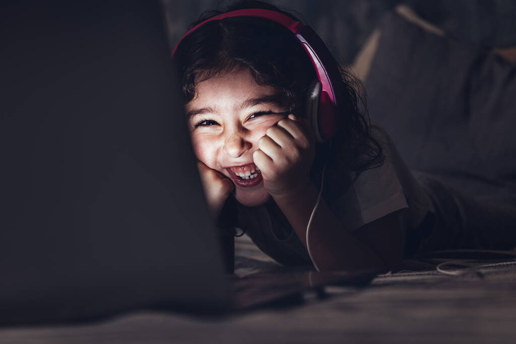 kleines Mädchen, das lachend im Bett liegt, während es sich in der Dunkelheit einen Film auf dem Laptop ansieht, hat ein rosafarbenes Headset, Kinder- und Technologiekonzept, Kopierplatz für Text - Foto, Bild