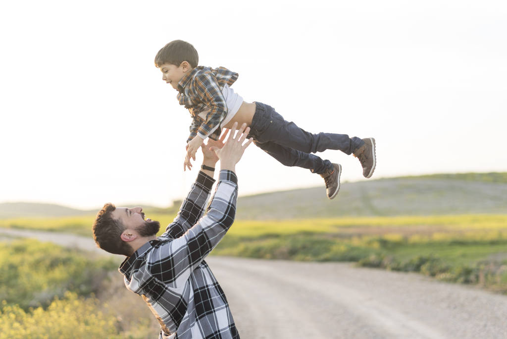 Ο πατέρας πετάει τον γιο του στον αέρα παίζοντας έξω, ευτυχισμένος την άνοιξη. - Φωτογραφία, εικόνα