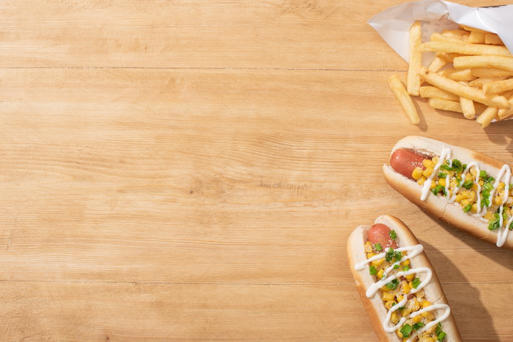 Blick von oben auf köstliche Hot Dogs mit Mais, grünen Zwiebeln und Mayonnaise in der Nähe von Pommes frites auf Holztisch - Foto, Bild