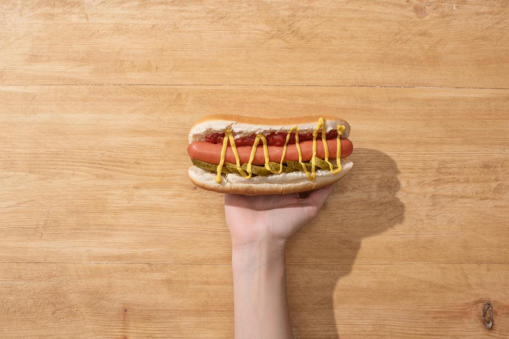 částečný pohled na ženu držící lahodný hot dog s nakládanými okurkami, hořčicí a kečupem na dřevěném stole - Fotografie, Obrázek