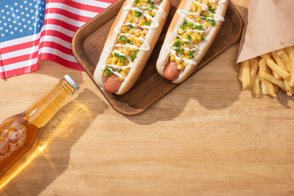 bovenaanzicht van heerlijke hotdogs met maïs, groene ui en mayonaise bij frietjes, Amerikaanse vlag en bier op houten tafel - Foto, afbeelding