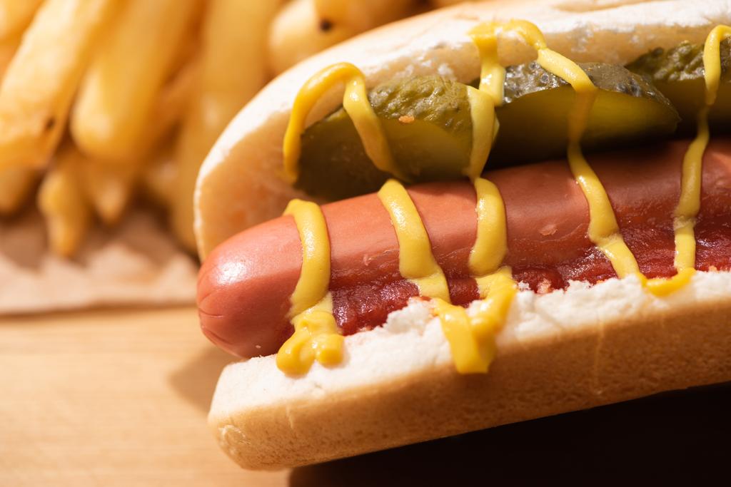 zblízka pohled na lahodný hot dog s nakládanými okurkami, kečupem, hořčicí u hranolků na dřevěném stole - Fotografie, Obrázek